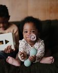 A baby sits on a couch holding a green mushie Silicone Baby Rattle Toy with a pacifier in their mouth, while an older child nearby plays with a shape-sorting toy. The softly lit background features wood paneling.