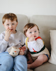 Two toddlers sit on a beige couch enjoying snacks. The older child, in a gray sweatshirt and blue pants, holds a mushie Snack Cup. The younger child wears a colorful outfit with a white shirt and looks at the camera.