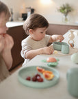 Two young children sit at a bright kitchen counter. One practices independent eating with mushie Silicone Toddler Starter Spoons and fruit, while the other plays with cups and a toy tea set beside plates of grapes and orange slices.