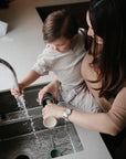 A woman helps a young child wash dishes at the kitchen sink using the mushie Silicone Baby Bottle Brush with silicone bristles, creating a warm and nurturing scene.