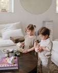 Two toddlers play in a cozy living room with cups and bowls as the mushie Snack Cup, a spill-proof silicone solution, sits nearby. Soft light highlights white sofas and neutral decor in the inviting space.