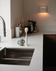 On a modern kitchen countertop with warm, cozy lighting sits a mushie Silicone Baby Bottle Brush, a green sippy cup with a straw, a sculpted candle, and a coffee maker in the background next to a stainless steel sink.
