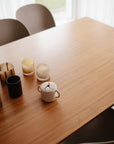 A wooden table holds five glass candle holders, a mushie Snack Cup for toddlers, and a small double-handled white cup. Four modern chairs surround the table as sunlight streams through the window in the background.