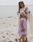A woman holds a baby in a mushie Organic Cotton Muslin Swaddle Blanket on a sandy beach.