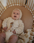 A smiling baby in a knitted bonnet and sweater lies in a cozy striped bassinet, cuddling the mushie Moon Lovey Blanket and holding a teething toy.
