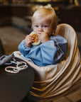 A blond-haired toddler sits in a high chair, wearing a mushie Long Sleeve Bib while eating a sandwich. On the table are a shark-shaped teething toy and several pacifier clips.