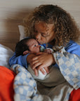 A young child with curly hair sits on a bed, lovingly holding and kissing a newborn wrapped in a mushie Organic Cotton Muslin Swaddle Blanket.