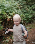 A young blonde-haired child in a striped shirt and backpack stands on a forest path, smiling while holding the mushie Silicone Baby Rattle Toy, enjoying a sensory experience with green foliage in the background.