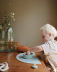 A young child with blonde hair sits at a wooden table, reaching into a bowl on a mushie Silicone Placemat. Soft natural light brightens the scene, with a vase of flowers, candle, and decorative items completing the cozy setting.