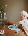 A young child with blonde hair sits at a wooden table, reaching into a small bowl with mushie Silicone Toddler Starter Spoons, while a tall vase with flowers and a lit candle sit nearby in the softly lit room.
