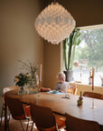 A young child practices self-feeding with mushie Silicone Toddler Starter Spoons at a wooden dining table, surrounded by plants, flowers, and gold candle holders beneath a large chandelier in a sunlit room.