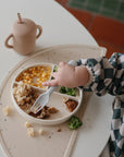 A young child in a checkered shirt uses a fork to eat pasta, corn, broccoli, and meat from a divided plate. Next to the plate are a tan sippy cup and a mushie Silicone Placemat made from food-grade silicone.