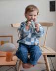 A smiling toddler in a blue checkered sweater and jeans sits barefoot on a wooden chair, playfully biting the mushie Robot Rattle Teether. A lamp, framed artwork, and small items are on the table beside them.