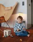 A young child sits on a tiled floor playing with wooden toys and a FRIGG Lucky Symmetrical Silicone Pacifier 2-Pack (6-18 Months) by FRIGG. A tan butterfly chair and table lamp create a cozy indoor scene in the background.