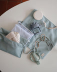 A neatly arranged flat lay displays mushie's Water Resistant Wet Bag, pastel blue pouches, a baby teether, pacifier clip, soft toy, and cloth—perfect for organized diaper bag storage—with a small sprig in a vase.