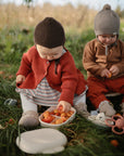Two young children sit on grass outdoors in warm clothes and hats. One reaches for apples from a mushie Lunchbox with built-in compartments, while the other holds fruit nearby. Toys and a plate are also visible.