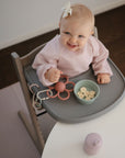 A smiling baby in a high chair wears a light pink bib with a bowl of sliced bananas and a pink flower-shaped teether on the tray, while the Mushie Splat Mat protects the floor beneath her.