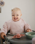 A smiling baby with a bow in her hair sits in a high chair, wearing the mushie Long Sleeve Bib in light pink. A green bowl and pink pacifier rest on the tray, all set against a softly lit, minimalistic background.