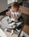 A baby sits in a high chair, wearing a bib, using the mushie Dinnerware Fork and Spoon Set to eat from a bowl of food. A soft toy rests on the tray as sunlight streams in through nearby windows.