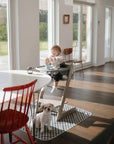 A baby in a high chair eats at a table in a bright, modern dining room. A small white dog stands on the Mushie Splat Mat near the high chair, looking at the camera as sunlight streams through large windows.
