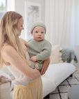 A woman with long blonde hair soothes her baby, who is wearing a light green knit outfit and hat, with the FRIGG Lucky Symmetrical Silicone Pacifier (6-18 Months) by FRIGG in a bright, cozy living room filled with natural light.