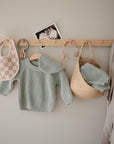 A light green knitted baby sweater, checkered bib, teether, photo, FRIGG Lucky Symmetrical Silicone Pacifier (6-18M), beaded pacifier clip, woven bag with clothes, and beige robe hang on a wooden wall rack against a light wall.