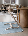 A Mushie Splat Mat lies under a modern wooden high chair next to a wooden dining table in a stylish, light-filled living and dining room with neutral decor and a sofa in the background.