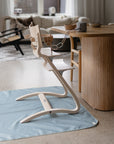 A modern, light wood high chair featuring a mushie Snack Cup sits on a blue mat in a cozy living-dining area with neutral furniture and large windows—perfect for toddlers.
