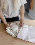 A person places folded baby clothes and a small white container into a white mushie Water Resistant Wet Bag on a wooden table, demonstrating convenient diaper bag storage with this practical, water-resistant accessory.