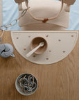 A wooden table displays a mushie Silicone Placemat in beige with rainbow designs, paired with a bowl, spoon, food-grade silicone linked rings, and a blue pacifier holder clipped to a wooden high chair.