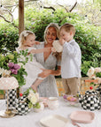 A woman and two kids pour tea from a Mushie Silicone Tea Play Set at a flower-decorated table.