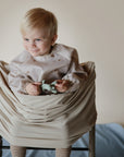 Smiling toddler in high chair holds toy, wearing mushie Multi-Use Cover and a light outfit.