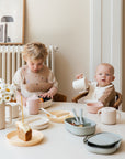 Two young children sit at a round table with cake, drinks, and pastel plates. One pours from a cup while the other eats from a non-toxic plastic mushie Lunchbox. Flowers and framed art decorate the bright, cozy room.