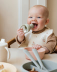 A smiling baby sits at a table, holding and chewing on the Mushie Silicone Fresh Food Feeder, with pastel bowls, utensils, and a cup in front—making self-feeding fun and easy.