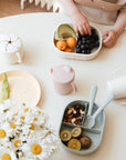 A toddler’s hand reaches for grapes in a lunchbox on a white table, surrounded by a mushie Snack Cup, cups, a plate, utensils, and a bouquet of daisies.