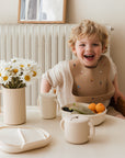 A smiling blond child wearing a bib sits at a table with a mushie Lunchbox, bowl of fruit, plate, cup, and vase of daisies in a bright, cozy room.