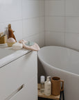 A minimalist bathroom features a white tub, white tiled walls, and a white vanity. Near the sink and on a wooden stool by the tub are toiletries, soap, and mushie Cradle Cap Brushes with silicone bristles.