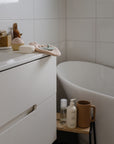 A minimalist bathroom with white tiled walls. A white cabinet holds toiletries and a cupcake-shaped soap. Nearby, a freestanding tub displays mushie Lavender Baby Shampoo & Body Wash beside other bath items on a wooden tray.
