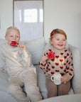 A young boy and girl sit on a light couch. The boy wears beige and holds a red heart lollipop, while the smiling girl in a red-heart sweater holds a mug. Nearby is her Mushie Bluey x Mushie Silicone Training Cup + Straw, made of food-grade silicone.
