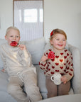 A young boy and girl sit on a light couch. The boy wears beige and holds a red heart lollipop, while the smiling girl in a red-heart sweater holds a mug. Nearby is her Mushie Bluey x Mushie Silicone Training Cup + Straw, made of food-grade silicone.