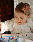 A young child with a pink bow smiles as she paints with watercolors, wearing a mushie Long Sleeve Bib to protect her clothes while holding a brush at the table in warm sunlight.