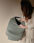 A woman with long hair, wearing beige, looks down at a baby car seat covered by the mushie Multi-Use Cover in sage green. The neutral background and minimal scene add to the calm atmosphere.