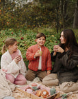 A woman and two children sit on a blanket outdoors, each holding a mushie Dinnerware Cup.