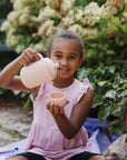 Smiling girl pours from Mushie Silicone Tea Play Set during an outdoor pretend tea party.