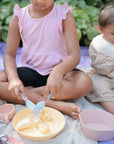 Two young children sit on a blanket outdoors. The older child in pink uses plastic utensils to cut a banana, while the younger in a bib looks at food in mushie Square Dinnerware Bowls. Pink plastic dinnerware set pieces are also nearby.