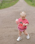 Toddler in pink wearing FRIGG Daisy Natural Rubber Pacifier, walking on a gravel path outside.