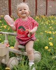 Smiling toddler with mushie Soft Spout Sippy Cup on tricycle, among yellow dandelions.