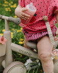 Toddler on a tricycle holding a mushie Soft Spout Sippy Cup, with a pacifier clipped on.