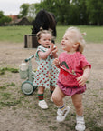 Two toddlers play outside, each holding a FRIGG Daisy Natural Rubber Pacifier 2-Pack.