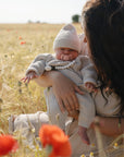 A woman holds a baby in a knitted outfit and hat among tall grass and red poppies. The calm baby gently sucks on the FRIGG Lucky Symmetrical Silicone Pacifier (0-6 Months), supporting oral development.